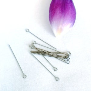 Può includere: Primo piano di un gruppo di spilli per la testa argentati con anelli alle estremità. Gli spilli sono sparsi su una superficie bianca, con un solo petalo di fiore viola sullo sfondo.