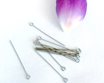 May include: A close-up of a group of silver head pins with loops on the ends. The pins are scattered on a white surface, with a single purple flower petal in the background.