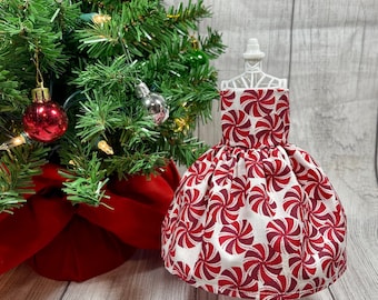 May include: A white dress with a red and white peppermint swirl pattern on a white mannequin. The dress is on a white stand in front of a Christmas tree.