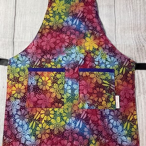 May include: A colorful apron with a floral pattern in shades of pink, blue, yellow, and green. The apron has two large pockets with purple trim. The apron is made of cotton fabric and has a tie closure.