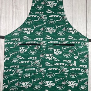 May include: Green and white kitchen apron with a repeating pattern of the New York Jets logo and text. The apron has three pockets.