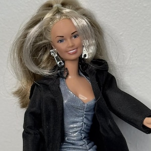 May include: A doll with long blonde hair styled in a high ponytail, wearing silver zipper-shaped earrings, a shimmering silver ruched top, and a black jacket. The doll has blue eyes, a light skin tone, and a neutral expression with a visible smile.