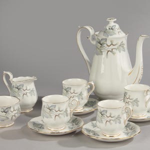Könnte beinhalten: Ein weißes Porzellan-Tee-Set mit einem zarten Blumendesign. Das Set beinhaltet eine Teekanne, eine Milchkännchen, eine Zuckerdose und vier Teetassen mit Untertassen.