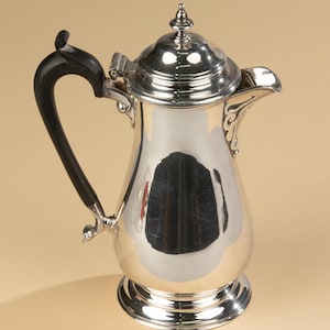 Op de afbeelding: Een zilveren koffiepot met een zwarte handgreep en een deksel. De pot heeft een gebogen tuit en staat op een ronde voet. De handgreep is gebogen en aan de zijkant bevestigd.