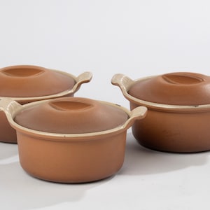 Puede incluir: Tres cazuelas de cerámica marrón con tapas. Las cazuelas tienen bordes y asas blancas.