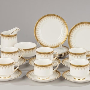 May include: A white porcelain tea set with gold detailing. The set includes cups, saucers, plates, a creamer, and a sugar bowl. The gold trim has a repeating pattern. The tea set is arranged on a light grey surface.