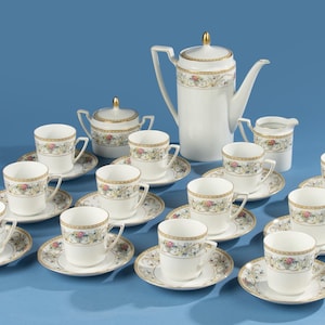 Puede incluir: Un juego de café de porcelana blanca con detalles florales y dorados. El juego incluye una cafetera, una jarra de crema, un azucarero y varias tazas y platillos. Las tazas y platillos están sobre un fondo azul.