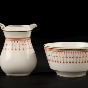 Puede incluir: Un juego de jarra de crema y azucarero de cerámica blanca. La jarra de crema tiene un asa y un pico, mientras que el cuenco es redondo. Ambas piezas presentan un patrón decorativo rojo y dorado alrededor del borde y el cuerpo. El conjunto está sobre un fondo negro.