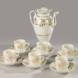 Puede incluir: Un juego de café de porcelana blanca con detalles dorados. El juego incluye una cafetera alta con tapa y seis tazas pequeñas con platillos a juego. El diseño de hoja de oro presenta patrones florales y de hojas.