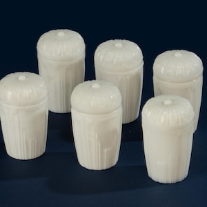 May include: Six white, lidded jars with a ribbed design on the sides and a floral pattern on the lids. The jars are arranged on a dark blue surface, showcasing their clean, classic aesthetic.