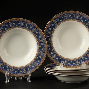 May include: A set of six white ceramic soup bowls featuring a wide rim decorated with a dark blue pattern of geometric shapes, small orange accents, and gold trim along the outer edge. One bowl is displayed upright on a clear stand.