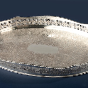 Könnte beinhalten: Ein ovales silberfarbenes Serviertablett mit einem komplizierten gravierten Blumenmuster auf der Basis, einer glatten ovalen Kartusche in der Mitte und einem erhöhten Galerie-Rand mit durchbrochenem Gitterdesign sowie integrierten Seitengriffen.