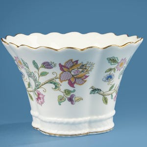 May include: White porcelain bowl with a scalloped rim and floral pattern. The bowl has a gold trim and a flared shape. The floral design includes purple, pink, yellow, and green elements, adding a touch of elegance.