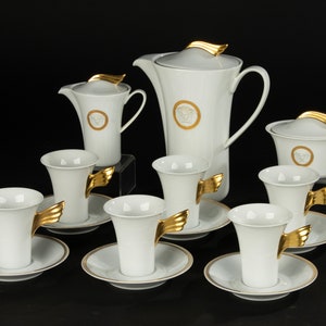 May include: A white porcelain tea set with gold accents. The set includes a teapot, creamer, sugar bowl, and six cups and saucers. Each piece features a gold winged handle and a gold medallion with a Medusa head design.