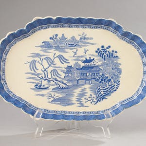 以下が含まれることがあります： 青と白の柳模様のデザインが施された楕円形のサービングプラッター。プラッターは、青いボーダーが付いたスカラップエッジを備えています。中央には、建物、木、川のある詳細な風景シーンが描かれています。