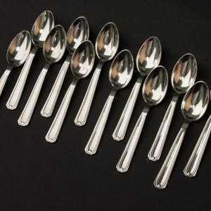 May include: A collection of twelve silver spoons arranged diagonally on a black surface. Each spoon has a rounded bowl and a handle with a decorative border. The spoons are reflective, showing the surrounding environment.
