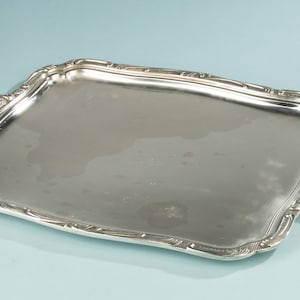 May include: A rectangular silver-colored serving tray with ornate handles and a decorative border. The tray has a reflective surface and is set against a light blue background. The handles have a scrolled design.