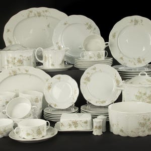 May include: A large white porcelain dinnerware set featuring a delicate light brown and green floral pattern. The collection includes various plates, bowls, lidded tureens, a gravy boat, a butter dish, and salt and pepper shakers with scalloped edges.