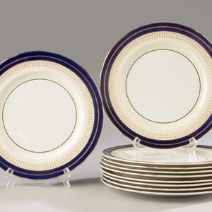 Puede incluir: Un juego de platos llanos redondos de cerámica con centro color crema, borde ancho azul marino y un patrón decorativo dorado en el borde. Los platos tienen bordes festoneados y se muestran tanto de pie como apilados en una columna.