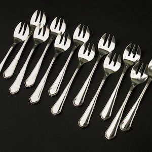 Pode incluir: Um conjunto de dez garfos prateados com três dentes cada, dispostos diagonalmente sobre uma superfície preta. Os garfos têm uma borda decorativa e ondulada nas suas pegas. A imagem mostra um conjunto de talheres elegantes.
