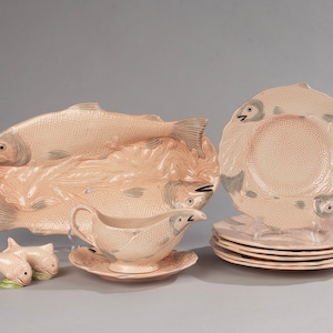 May include: A set of salmon-colored ceramic serving dishes featuring a fish design. The set includes a large oval platter, a gravy boat with a matching saucer, and five smaller plates. The dishes are all decorated with a raised fish design and have a textured surface.
