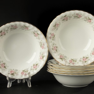 May include: A set of white ceramic bowls featuring a scalloped edge with a gold trim. The interior rim of each bowl is decorated with a repeating pattern of small pink roses and green leaves. One bowl is displayed upright on a clear stand.