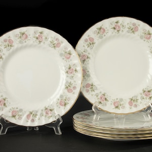 May include: A set of six white ceramic plates featuring a scalloped edge with a gold trim. The rim of each plate is decorated with a repeating pattern of small pink roses and light blue floral accents against a white background.