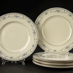 Op de afbeelding: Een set ronde keramische dinerborden met een gebroken wit midden en een decoratieve rand met een blauw bloemmotief en een dunne zilveren rand. Eén bord staat rechtop op een doorzichtige plastic standaard, vijf andere liggen gestapeld.