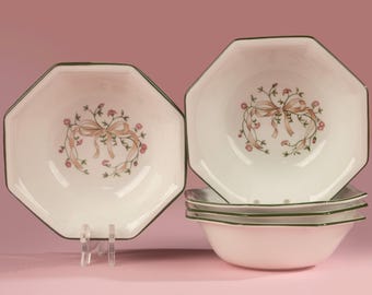 May include: A set of white octagonal bowls featuring a central floral motif with pink flowers, green leaves, and a tan ribbon. Each bowl has a dark green rim. One bowl is displayed upright on a clear stand, while three others are stacked below.