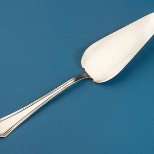 Könnte beinhalten: Ein silberfarbener Tortenheber mit einer breiten, dreieckigen Klinge mit glatter, polierter Oberfläche und einem schlanken, länglichen Griff. Der Griff zeigt ein dezentes, konisches Design mit einem dekorativen Schnörkel am Ende.