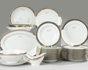 May include: A collection of white porcelain dinnerware with gold and silver trim. The set includes plates, bowls, serving dishes, and a gravy boat. Each piece is adorned with a silver monogram. A complete set for elegant dining.