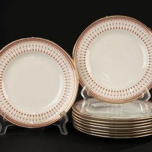 May include: A collection of white porcelain plates with a red and gold patterned border. The plates have a gold rim and are displayed on clear stands. The design features a repeating arrow pattern.