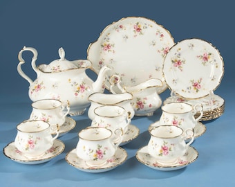 Puede incluir: Un juego de té de porcelana con base blanca, decorado con motivos florales de rosas rosas y amarillas y bordes dorados. El conjunto incluye una tetera, una lechera, un azucarero, una bandeja grande y seis tazas con sus platos a juego.