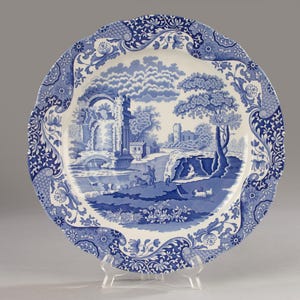 Può includere: Un piatto rotondo in ceramica con bordo smerlato caratterizzato da un motivo blu e bianco. La scena centrale raffigura un paesaggio con rovine in pietra, alberi e figure vicino a un fiume, mentre il bordo presenta motivi floreali.