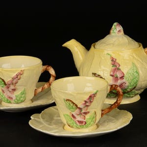 Könnte beinhalten: Ein Vintage-Tee-Set mit einer gelben Teekanne, zwei Teetassen und Untertassen. Die Teekanne und die Tassen haben ein florales Design mit rosa und grünen Blumen.