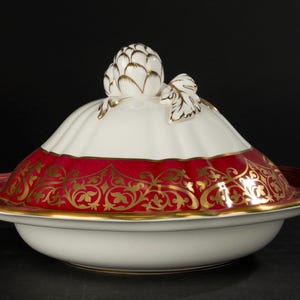 Può includere: Un piatto da portata in porcellana bianca con un bordo rosso e dorato e un motivo floreale decorativo. Il piatto ha un coperchio con un pomo a forma di carciofo dorato e bianco.