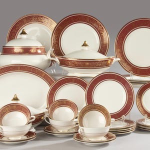 May include: A dinnerware set featuring cream-coloured porcelain with wide, deep red borders and intricate gold filigree patterns. The collection includes lidded serving tureens, dinner plates, salad plates, and teacups with matching saucers.