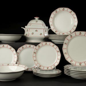 Op de afbeelding: Een wit keramisch servies met een rood en groen bessenpatroon. De set bevat borden, kommen en een serveerschaal met deksel. De borden hebben een geschulpte rand en een rode rand met bessenaccenten.