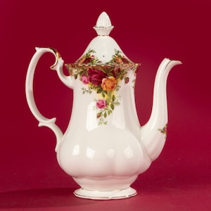 Puede incluir: Tetera de porcelana blanca con un patrón floral de rosas rojas, amarillas y rosas. La tetera tiene un borde dorado y una tapa con un pomo.