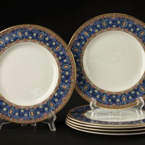 May include: A set of six white ceramic plates featuring a wide border with a dark blue background, intricate geometric patterns in shades of light blue, pink, and gold, and a thin gold rim along the outer edge of each circular dinner plate.