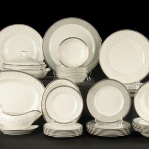 Op de afbeelding: Een grote serviesset bestaande uit off-white keramische delen met een delicaat grijs krulpatroon langs de randen. De collectie bevat diverse borden, kommen, een terrine met deksel en een sauskom, allemaal in een gelaagde opstelling.
