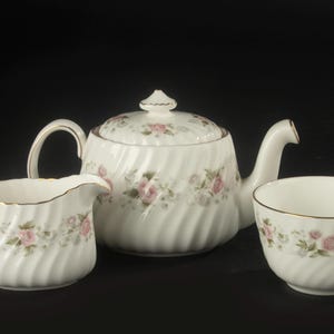May include: A white porcelain tea set featuring a teapot with a lid, a creamer, and a sugar bowl. Each piece has a swirling ribbed texture, a delicate floral pattern of pink roses and green leaves, and thin gold trim along the edges.