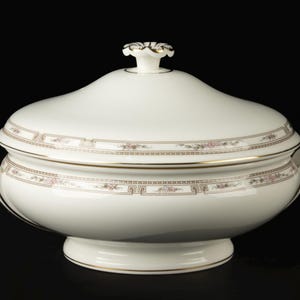 Puede incluir: Una fuente de porcelana blanca con tapa. El plato tiene un borde decorativo con ribetes dorados y detalles florales. La tapa está rematada con un asa decorativa. El plato está sobre un fondo negro.