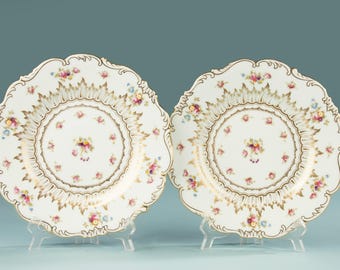 May include: Two white porcelain plates with scalloped edges and floral designs. The plates feature a gold rim and a central floral arrangement with pink, yellow, and blue flowers. The plates are displayed on clear stands.