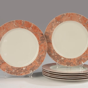 Op de afbeelding: Een set van zes ronde dinerborden met een effen crèmekleurig midden en een brede rand versierd met een gemarmerd patroon in tinten terracotta, oranje en grijs. De borden zijn gerangschikt met één rechtopstaand bord en vijf gestapelde borden.