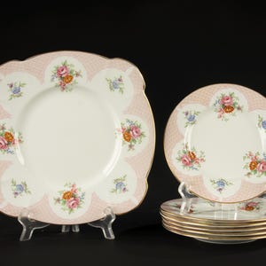 May include: A set of vintage porcelain plates with a floral design. The plates have a pink scalloped border with a repeating pattern and gold trim. The center of the plates is white, with colorful floral bouquets.