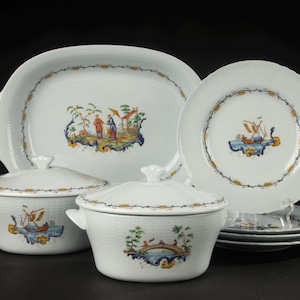 Op de afbeelding: Een wit porseleinen servies met een textuur van vlechtwerk en handgeschilderde scènes van figuren en boten. De set bevat een grote ovale schaal, twee serveerschalen met deksel en handvatten, en een stapel borden.