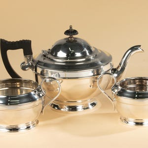 May include: A silver tea set with a teapot, creamer, and sugar bowl. The teapot has a black handle and a decorative finial. The creamer and sugar bowl have curved handles. The set is displayed on a light brown surface.
