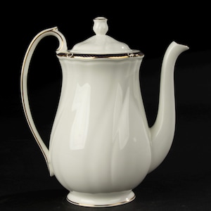 Pode incluir: Uma cafeteira de porcelana branca com uma alça e bico curvos. A cafeteira tem uma tampa com um pequeno botão e é adornada com ouro e uma faixa azul escura. A base também é adornada com ouro.