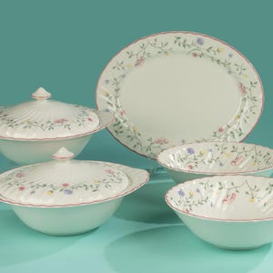 Pode incluir: Um conjunto de jantar em porcelana branca com padrões florais. O conjunto inclui duas tigelas de servir com tampa, duas tigelas menores e um prato oval. Cada peça tem uma borda ondulada e é decorada com flores rosa, azuis e amarelas.